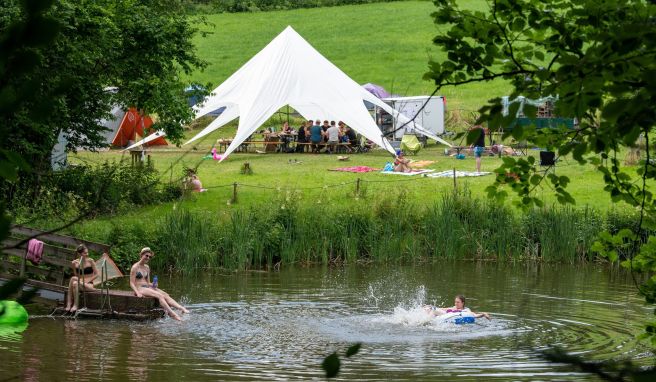 Teilnehmer eines Ferienlagers schwimmen in einem See, während die anderen zusammen im Gemeinschaftszelt sitzen.